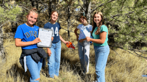 Four young women stand in a grassy, tree-filled outdoor area, holding clipboards and a small tool, smiling at the camera.